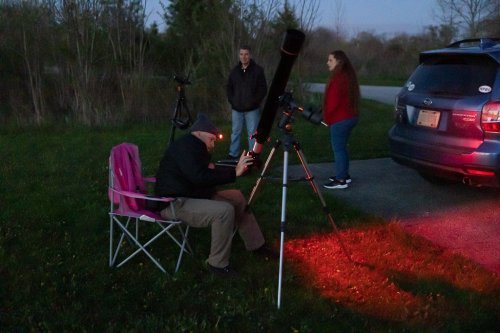 A group of three people stargazing at dusk, with telescopes set up and a car nearby. One person sits in a chair.