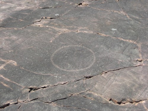 Petroglyph on rock surface featuring a circular design, surrounded by cracks and natural textures.