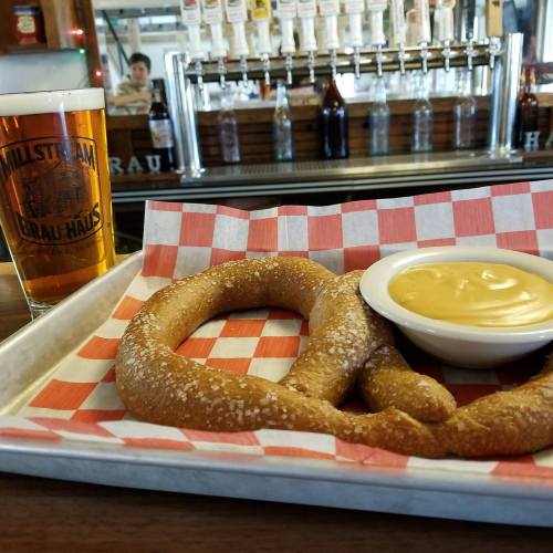 A soft pretzel with cheese dip on a checkered tray, alongside a pint of beer in a bar setting.