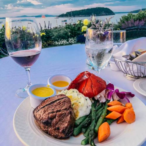 A plate of steak, lobster, vegetables, and sauces, with wine and water glasses, set against a scenic waterfront view.