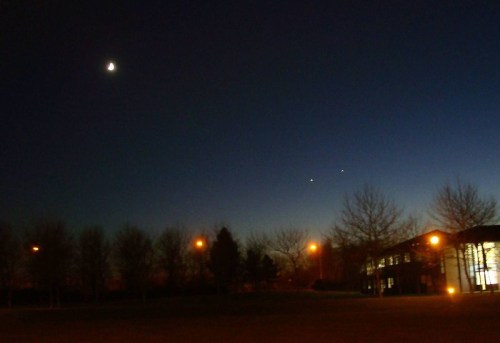 A crescent moon and two bright planets shine in a twilight sky above silhouetted trees and a building.