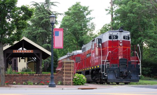 A vintage red train parked near a small station with a flag and greenery in Lebanon.
