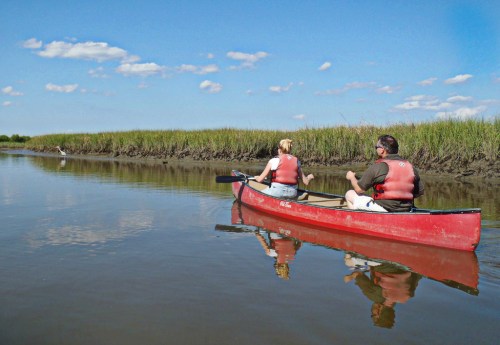 Two people in a red canoe paddle along a calm river surrounded by tall grass and a clear blue sky.