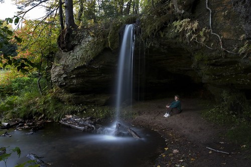 A serene scene of a person sitting by a waterfall in a lush, green forest. Water cascades over rocks into a calm pool.