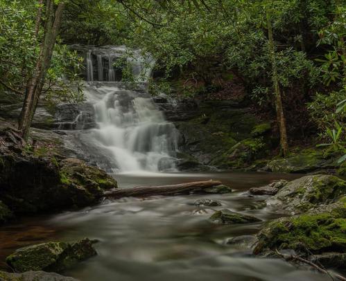 A serene waterfall cascades over rocks, surrounded by lush greenery and a calm pool below.