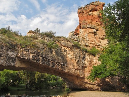A natural stone arch bridge over a calm river, surrounded by lush greenery and rocky cliffs under a blue sky.