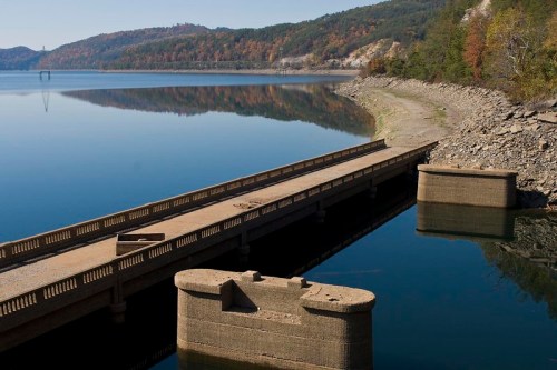 A serene lake scene with a concrete pier extending into calm waters, surrounded by autumn-colored hills.