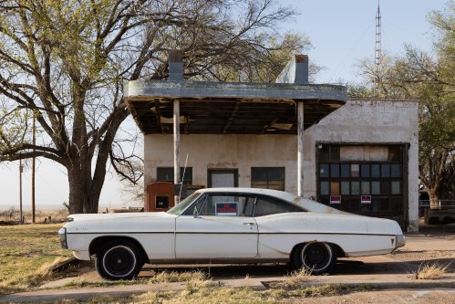 A vintage white car parked beside an old, abandoned gas station surrounded by trees and open land.