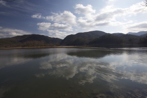 A serene lake reflects clouds and mountains under a blue sky, surrounded by lush greenery.