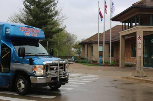 A blue shuttle bus stops at a crosswalk near a building with flags and a sign.