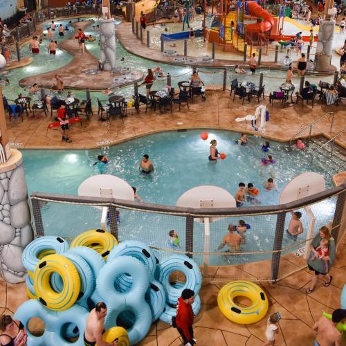 A busy indoor water park with pools, slides, and families enjoying the attractions. Colorful inflatable tubes are stacked nearby.