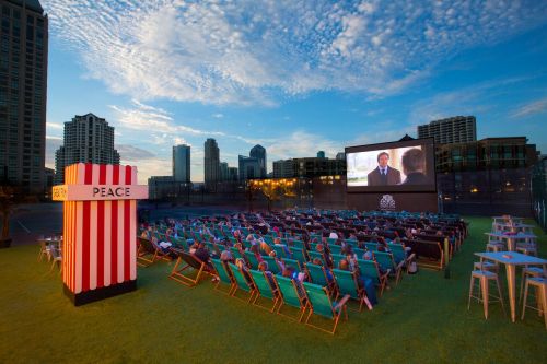 Outdoor movie screening with a large screen, colorful chairs, and a popcorn stand, set against a city skyline at dusk.