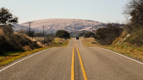 A long, straight road leads toward a large, rocky hill under a clear blue sky, surrounded by greenery.