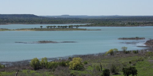 A serene landscape featuring a calm blue lake surrounded by greenery and distant hills under a clear sky.
