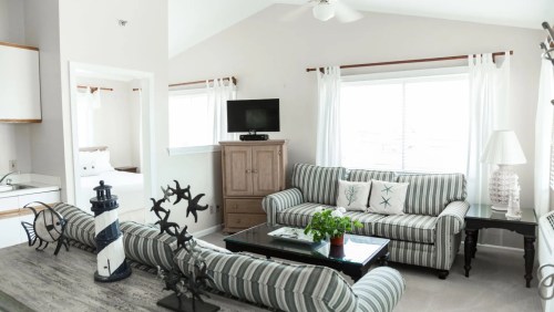 Bright, airy living room with striped sofas, a coffee table, and nautical decor, featuring a TV and large windows.