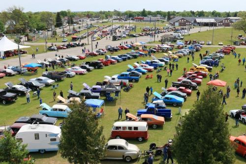 Aerial view of a car show featuring a variety of classic cars parked on a grassy area with spectators.