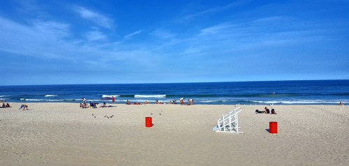A sunny beach scene with people relaxing on the sand, gentle waves, and a clear blue sky. Red barrels are visible.