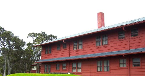 A red wooden building with a chimney, surrounded by greenery and trees under a cloudy sky.