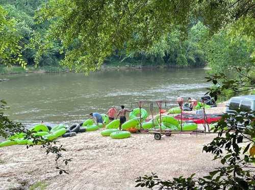 People preparing green inflatable tubes by a riverbank surrounded by trees. A vehicle is parked nearby.
