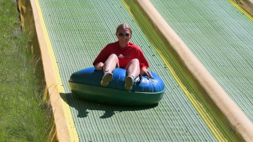 A person in a red shirt and sunglasses slides down a green water slide on a blue inflatable tube.