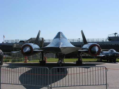 A black jet aircraft on display, viewed from the front, with a clear blue sky in the background and barriers in the foreground.