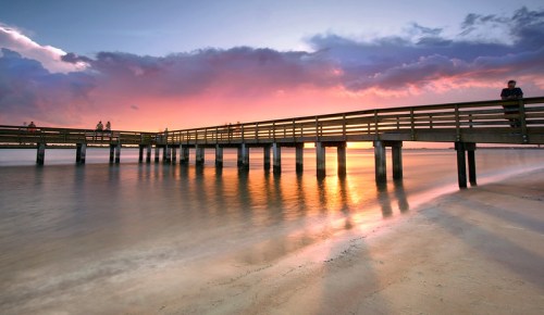 A wooden pier extends into calm waters at sunset, with vibrant clouds reflecting in the water.