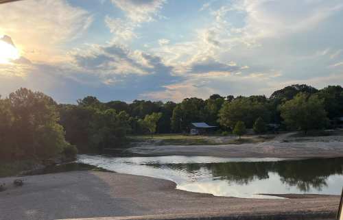 A serene river scene at sunset, with trees lining the banks and a small cabin in the background.