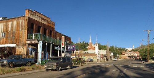 A small town street with rustic buildings, a café, and church steeples in the background under a clear blue sky.