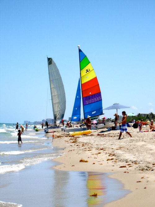 Two colorful sailboats on a sunny beach, with people enjoying the water and sand under a clear blue sky.