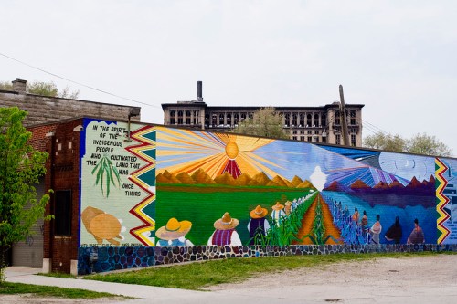 Colorful mural depicting indigenous themes, mountains, and people working the land, with a historic building in the background.