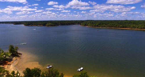 A serene lake surrounded by lush greenery and blue skies, with boats anchored near the sandy shore.