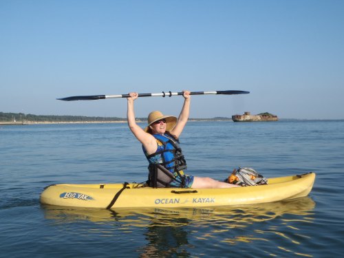 A person in a yellow kayak raises a paddle above their head, smiling against a clear blue sky and calm water.