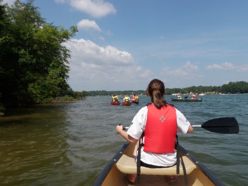 A person in a kayak paddles on a lake, with other kayakers and canoers visible in the background under a blue sky.
