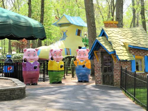 Three large pig statues in colorful outfits stand near a whimsical house and a giant shoe structure in a park.