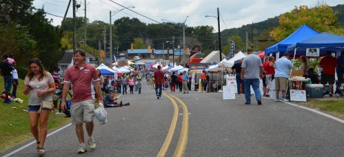 A lively street festival with vendors under blue tents, people walking, and colorful decorations in the background.