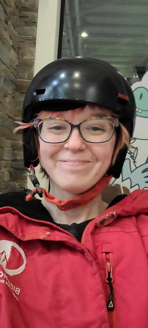 A person wearing a black helmet and red jacket smiles for the camera, with a cartoon character in the background.