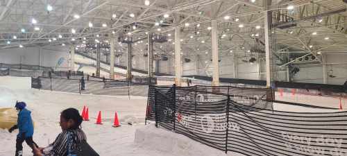 Indoor snow facility with ski slopes, safety barriers, and orange cones. People are enjoying winter sports activities.