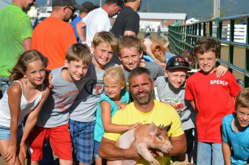A group of children and an adult pose together, smiling, with a pig in a farm setting.