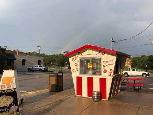 A colorful popcorn stand with a rainbow in the background, set in a parking lot with a few cars nearby.