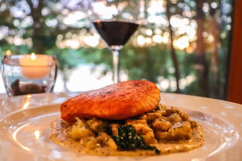 A plate of salmon on a bed of pasta, with a candle and a cocktail in the background, set against a scenic view.