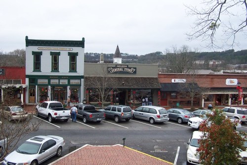 A view of a small town street with shops, parked cars, and a cloudy sky in the background.