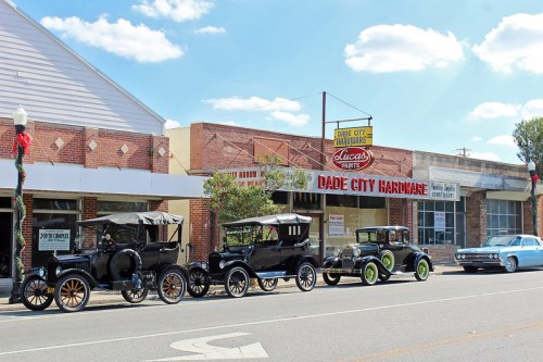 A row of vintage cars parked in front of a hardware store on a sunny day, with festive decorations visible.