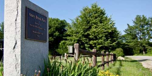 Sign for Amey Brook Park, surrounded by greenery and a wooden fence, with a clear blue sky in the background.