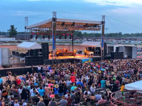 A lively outdoor concert with a large crowd enjoying a performance on stage at sunset.