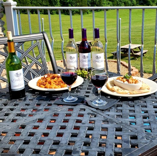 A table set with wine bottles, glasses, and plates of food, surrounded by green fields in the background.