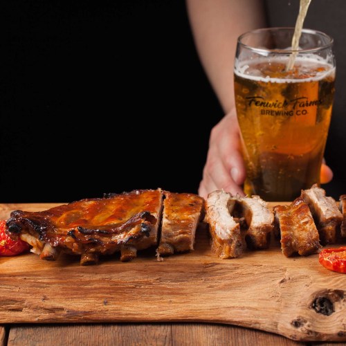 A wooden platter with sliced grilled meat and a glass of beer in the background.