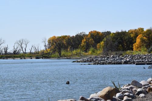 A serene lake scene with rocky shores and trees displaying autumn colors under a clear blue sky.