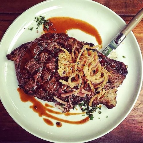 A juicy steak topped with crispy onions, served on a white plate with a knife, garnished with herbs and sauce.