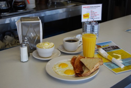 A diner breakfast featuring two eggs, bacon, toast, coffee, orange juice, and a bowl of oatmeal on a table.