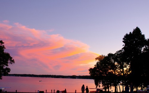 A serene lakeside scene at sunset, with vibrant pink and purple clouds reflecting over the water and silhouettes of trees.